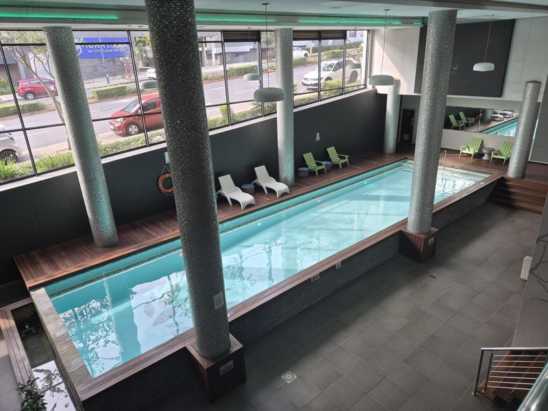 Indoor lap pool area featuring mosaic-tiled support columns, timber decking surround, and large floor-to-ceiling windows overlooking the street.