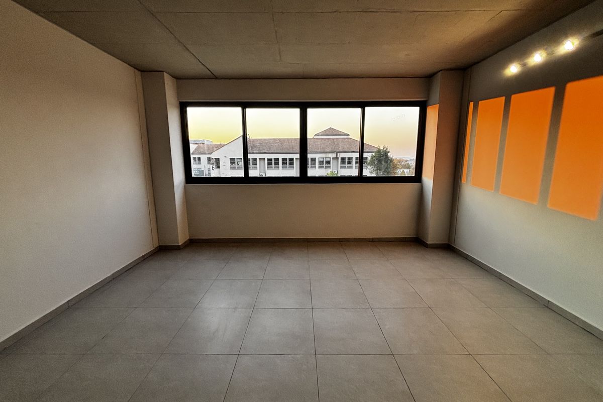A bright, empty living area featuring large windows, neutral tiled flooring, and modern track lighting, offering a clean and versatile space.