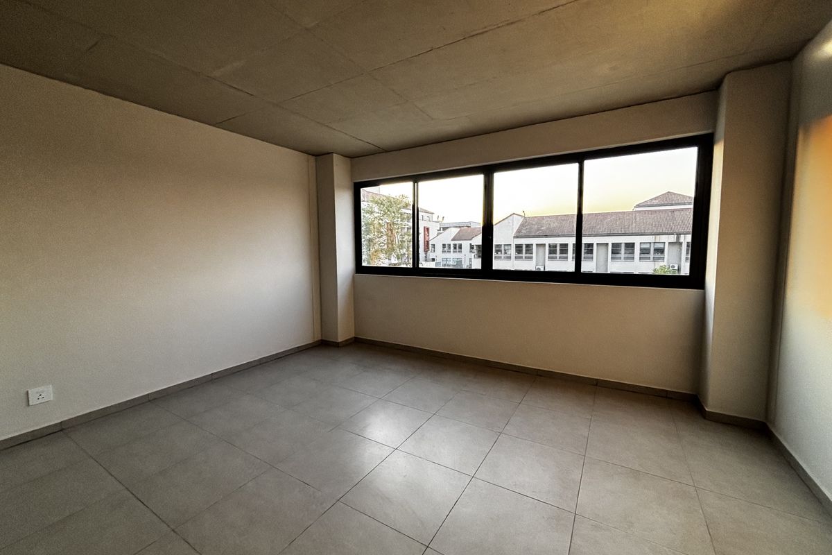 A bright, empty bedroom featuring neutral floor tiles, a raw concrete-style ceiling, and large windows offering an urban view.
