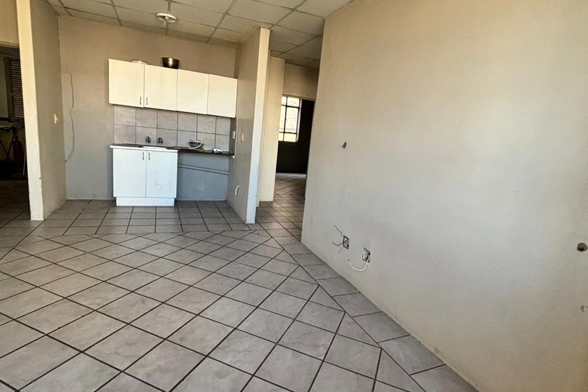 An open-plan living and kitchen area featuring tiled flooring, a drop ceiling, and basic built-in kitchen cabinetry in need of renovation.
