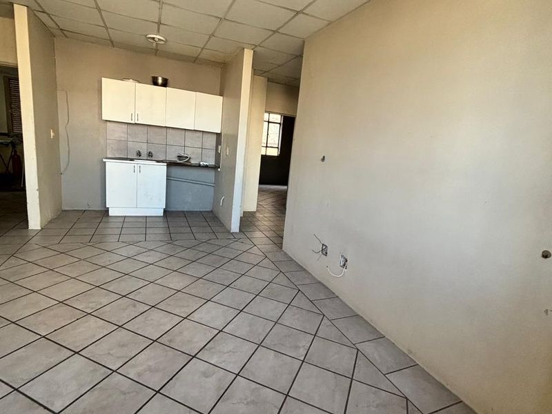 An open-plan living and kitchen area featuring tiled flooring, a drop ceiling, and basic built-in kitchen cabinetry in need of renovation.
