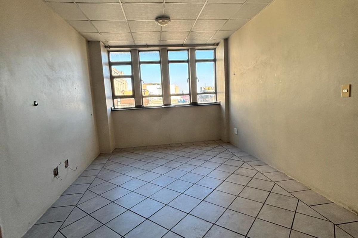 An empty, dated room featuring tiled flooring, a drop ceiling, and large windows providing natural light and an urban view.