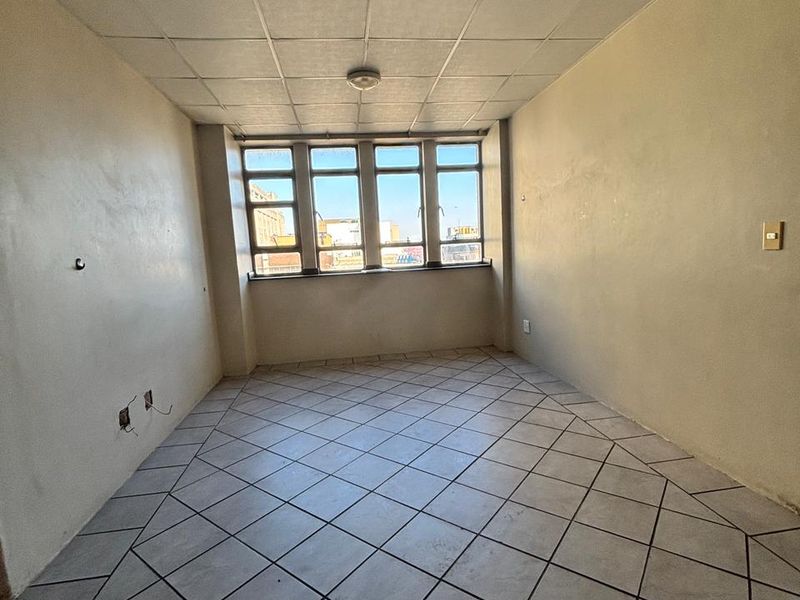 An empty, dated room featuring tiled flooring, a drop ceiling, and large windows providing natural light and an urban view.