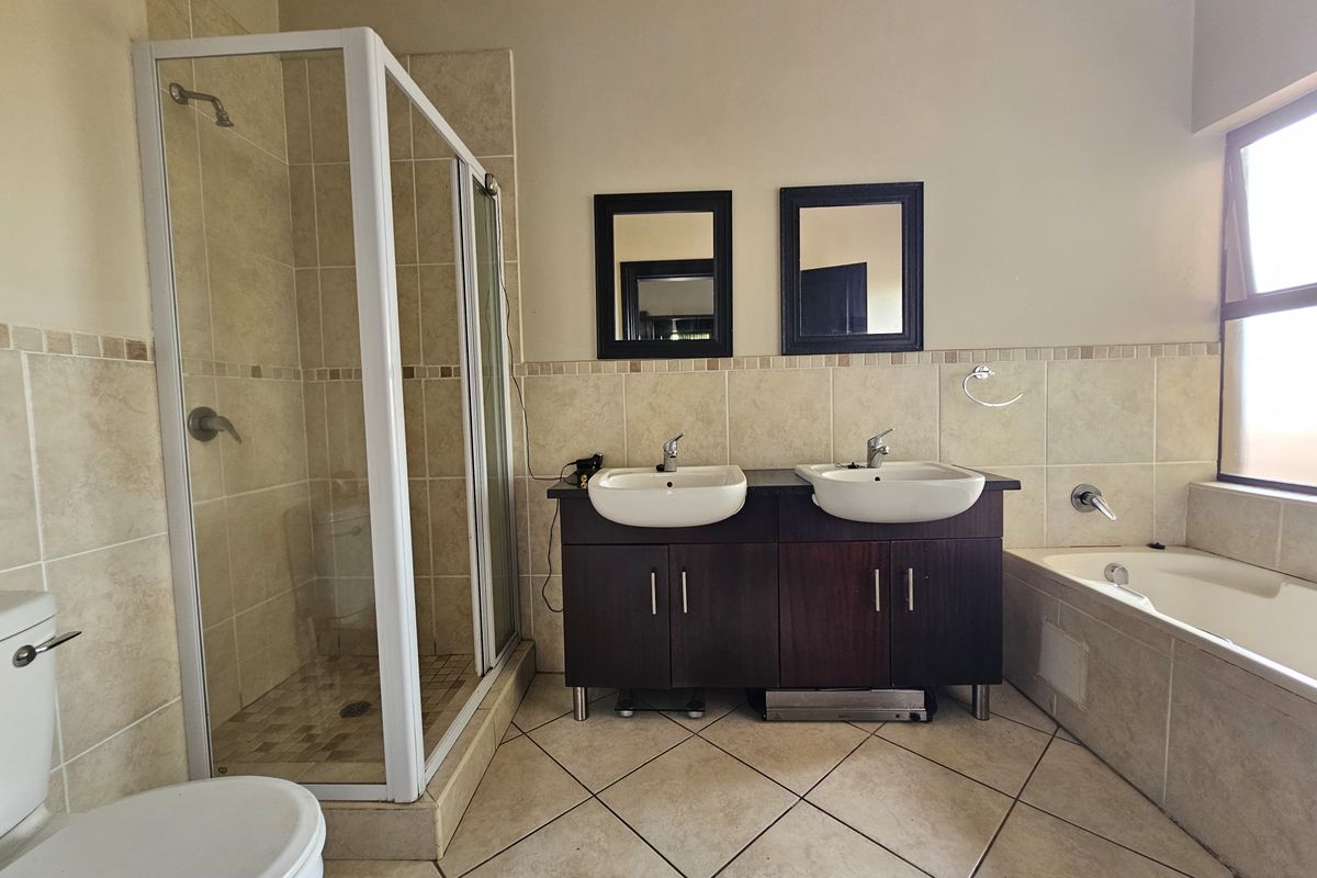 A functional bathroom featuring a double vanity with dark cabinetry, a glass-enclosed shower, a bathtub, and neutral floor-to-ceiling wall tiling.