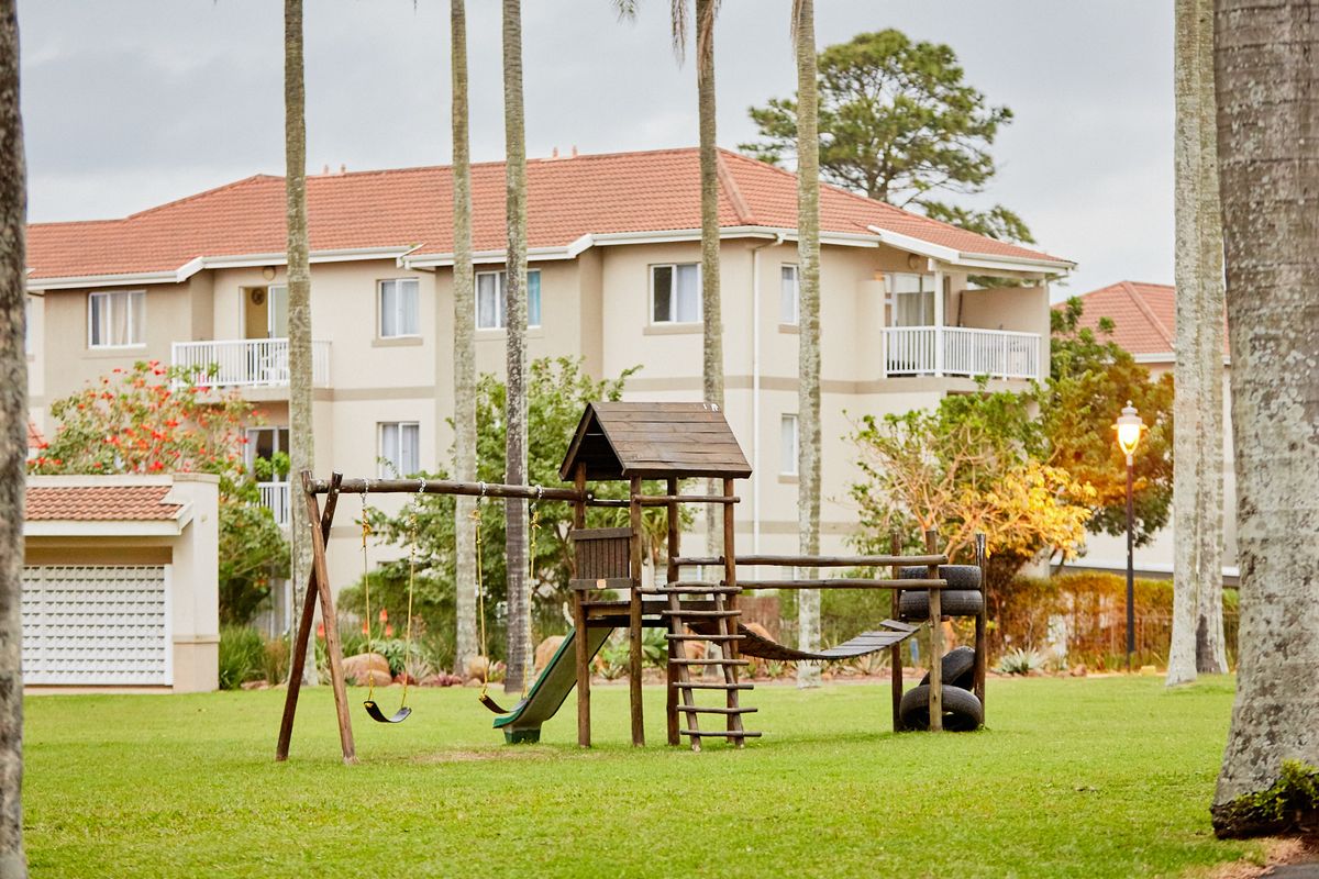 A view of the apartment complex exterior featuring a landscaped garden area with a wooden playground structure and mature palm trees.