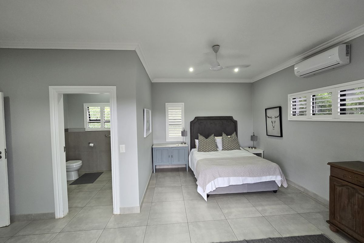 Spacious bedroom featuring neutral floor tiles, plantation shutters, a ceiling fan, and a wall-mounted air conditioning unit, with an adjoining en-suite bathroom.
