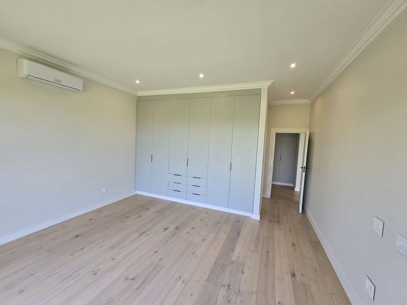 A bright, modern bedroom featuring light wood flooring, recessed ceiling lighting, built-in floor-to-ceiling cabinetry, and a wall-mounted air conditioning unit.