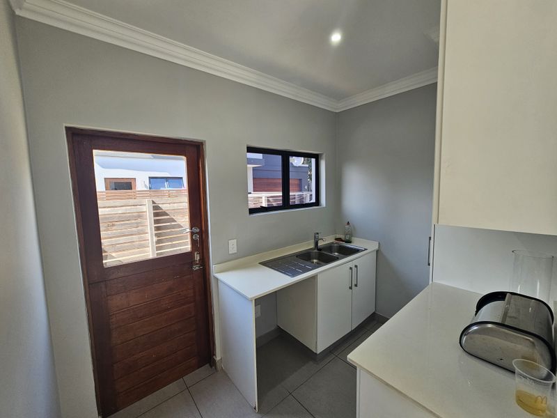 A functional scullery area featuring a stainless steel double sink, white cabinetry, tiled flooring, and direct access to the exterior via a wooden door.