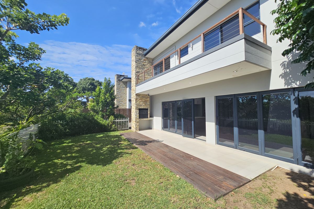 A modern rear exterior view featuring a spacious patio, built-in stone fireplace, timber decking, and large glass sliding doors leading to the interior.