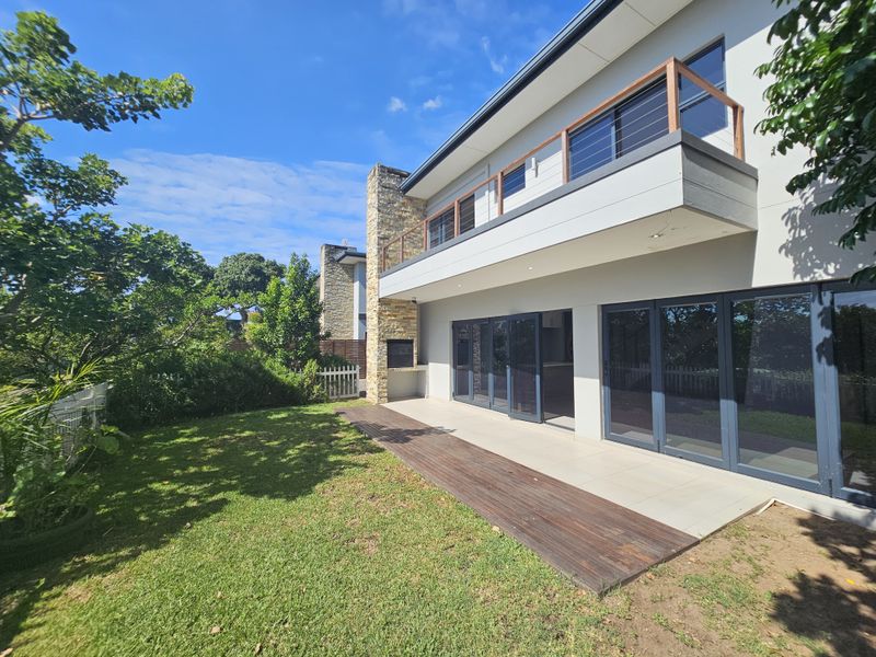 A modern rear exterior view featuring a spacious patio, built-in stone fireplace, timber decking, and large glass sliding doors leading to the interior.