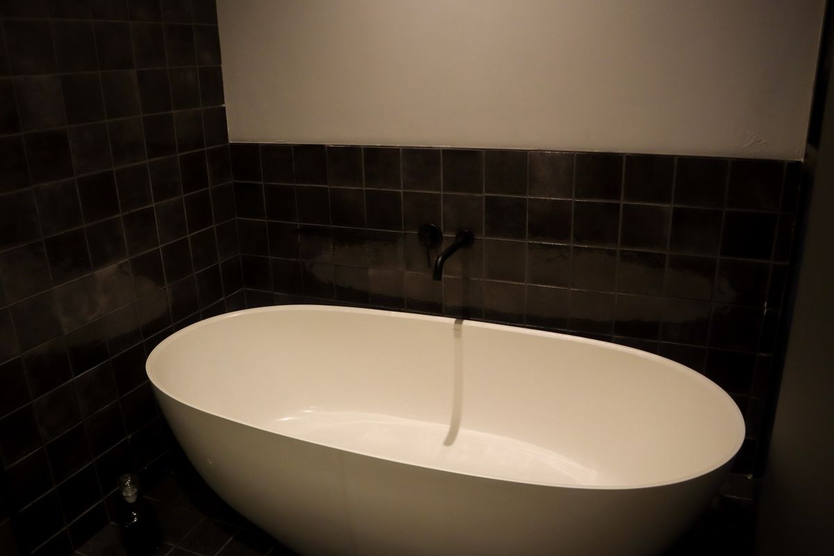 A modern bathroom featuring a freestanding white bathtub set against dark, square-tiled walls with matte black wall-mounted faucet fixtures.