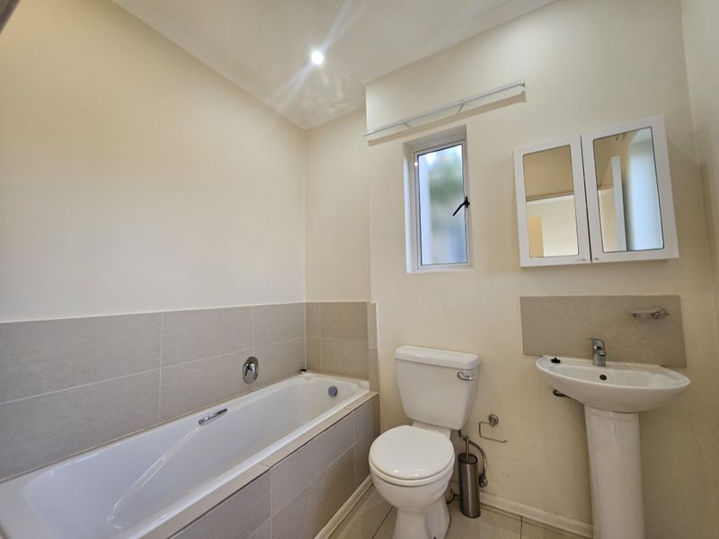A clean, functional bathroom featuring a built-in bathtub, pedestal basin, toilet, and wall-mounted medicine cabinet with neutral tiling.