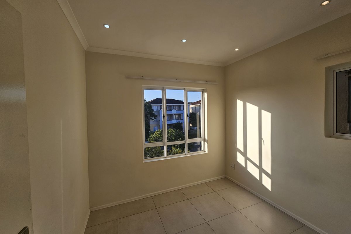 A bright, empty bedroom featuring neutral tiled flooring, recessed ceiling lighting, and large windows providing natural light and neighborhood views.
