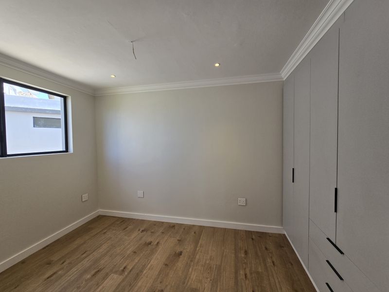 A bright, modern bedroom featuring light wood-look flooring, neutral walls, recessed ceiling lighting, and extensive built-in cabinetry with sleek black hardware.