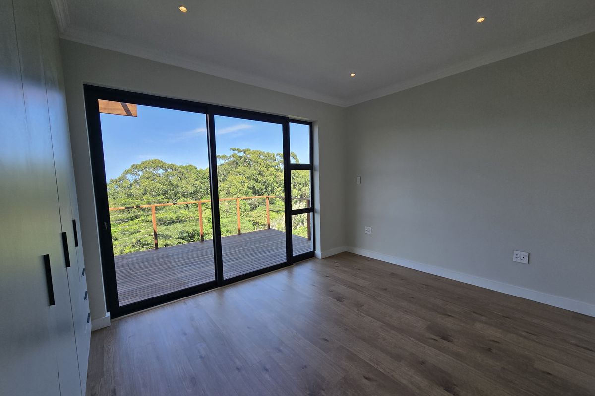 A bright, modern bedroom featuring wood-look flooring, built-in cupboards, recessed ceiling lighting, and large sliding glass doors opening onto a wooden deck with treetop views.