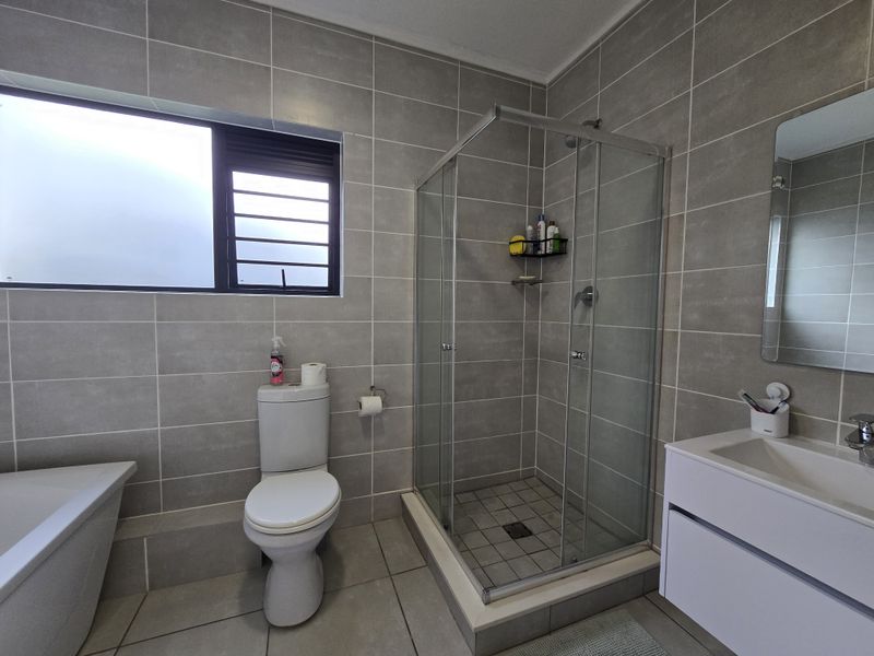 Modern bathroom featuring full-height grey wall tiling, a glass-enclosed shower, a white vanity unit, and a separate bathtub.