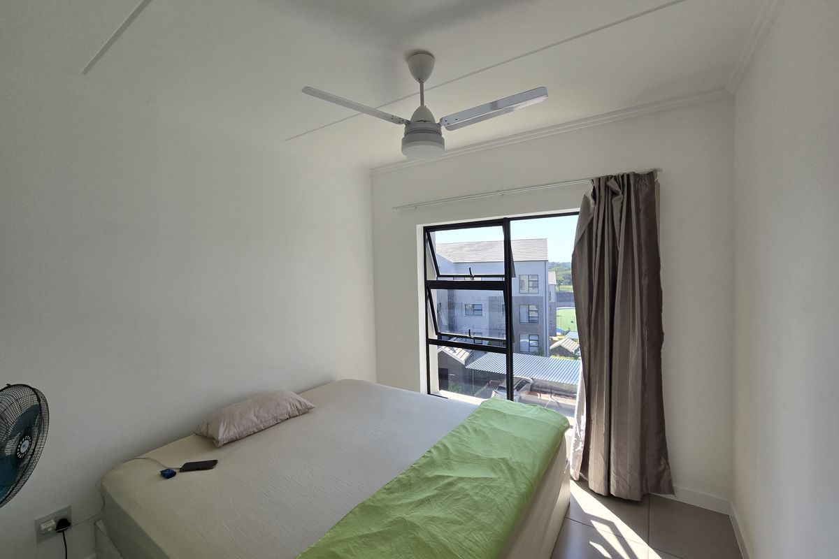 A bright bedroom featuring tiled flooring, a ceiling fan, and a large window providing natural light and views of the surrounding complex.