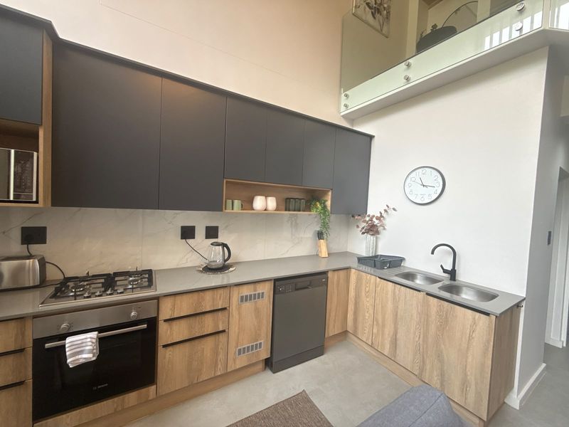Modern kitchen featuring sleek dark and wood-tone cabinetry, stone countertops, integrated appliances, and a double sink, set beneath a mezzanine level with glass balustrading.
