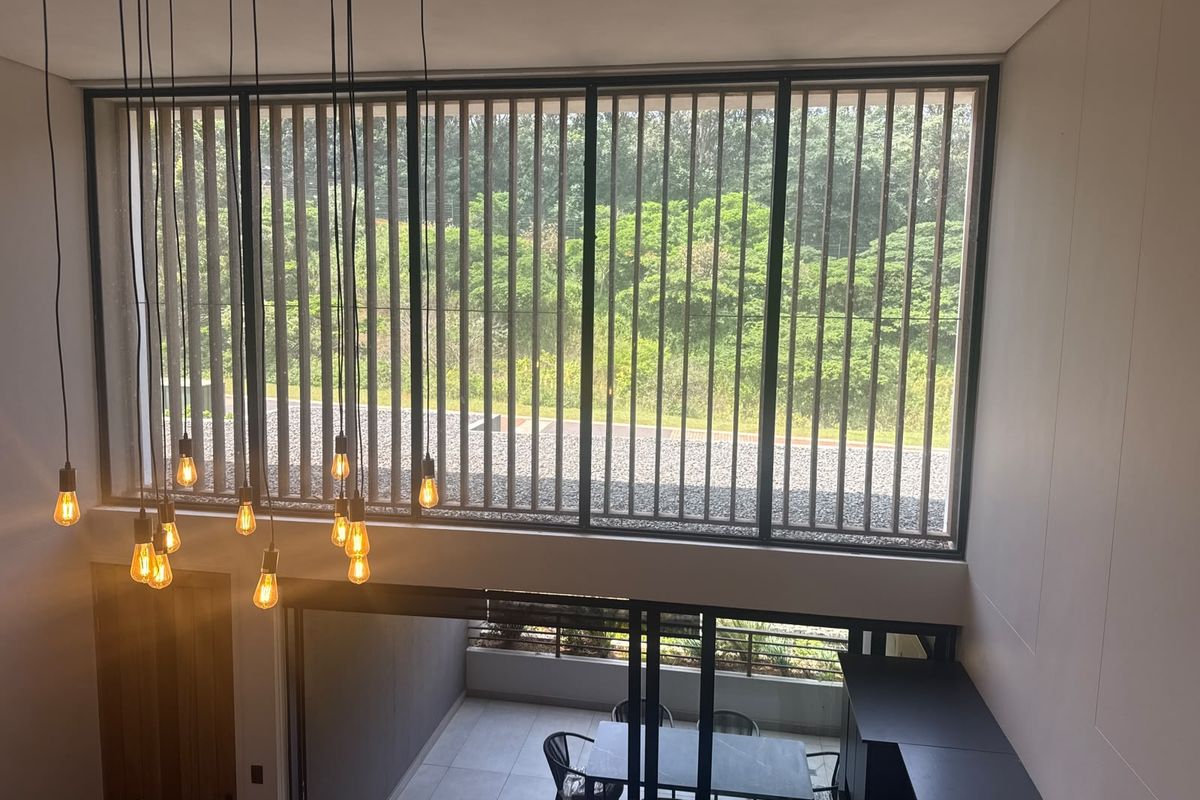 Spacious double-volume living area featuring large floor-to-ceiling windows with integrated timber privacy screens and modern pendant lighting, overlooking an outdoor patio space.