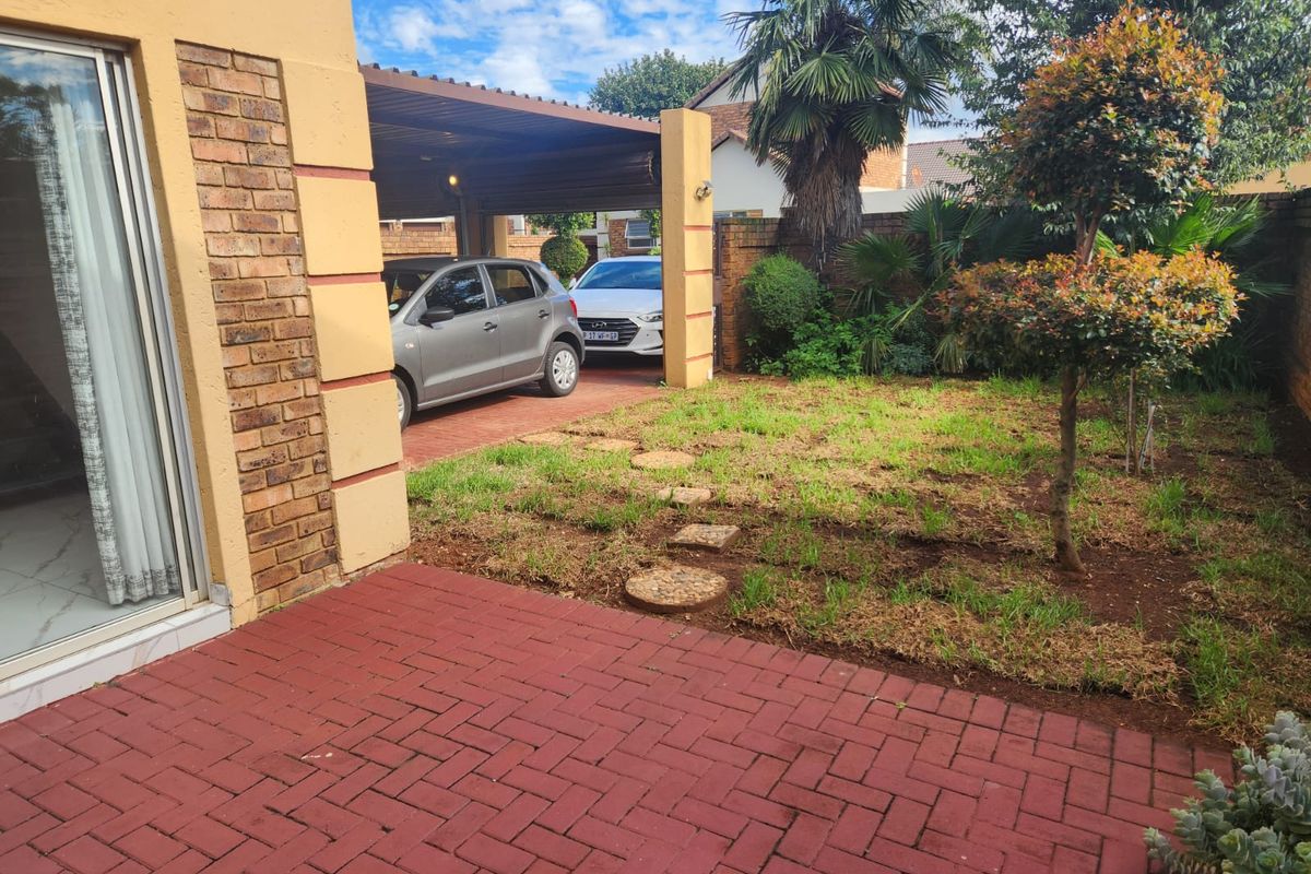 A rear garden area featuring a red brick paved patio, a covered carport, and a lawn space with established trees.