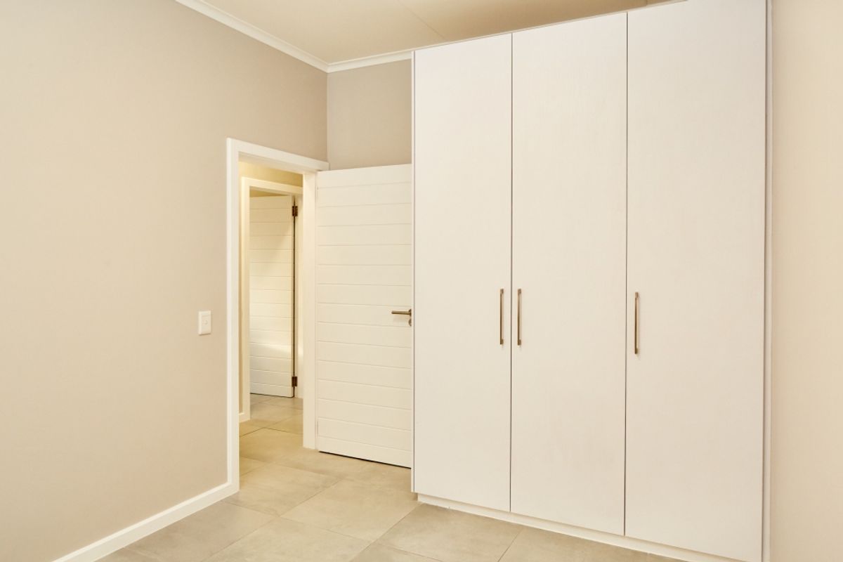 A clean, neutral bedroom featuring modern built-in wardrobes, tiled flooring, and access to an adjoining hallway.