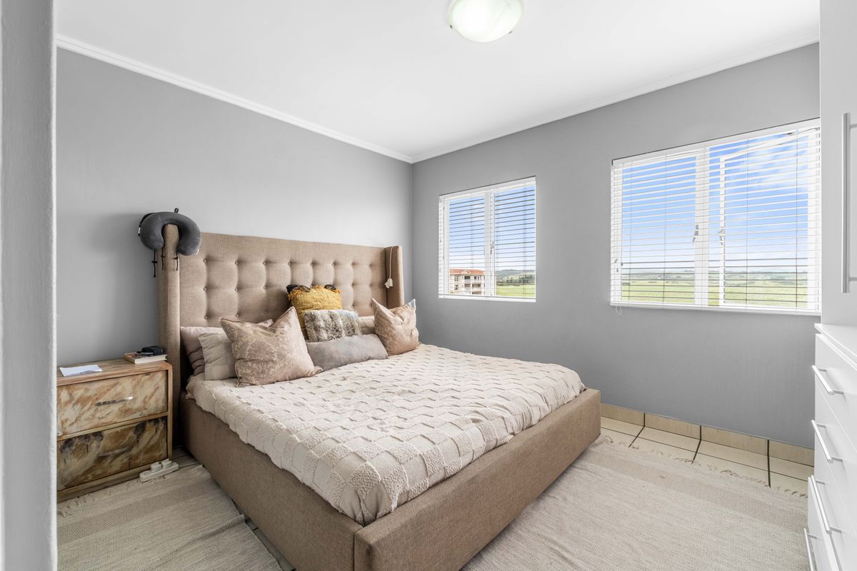 A bright bedroom featuring neutral grey walls, dual windows with venetian blinds, and tiled flooring.
