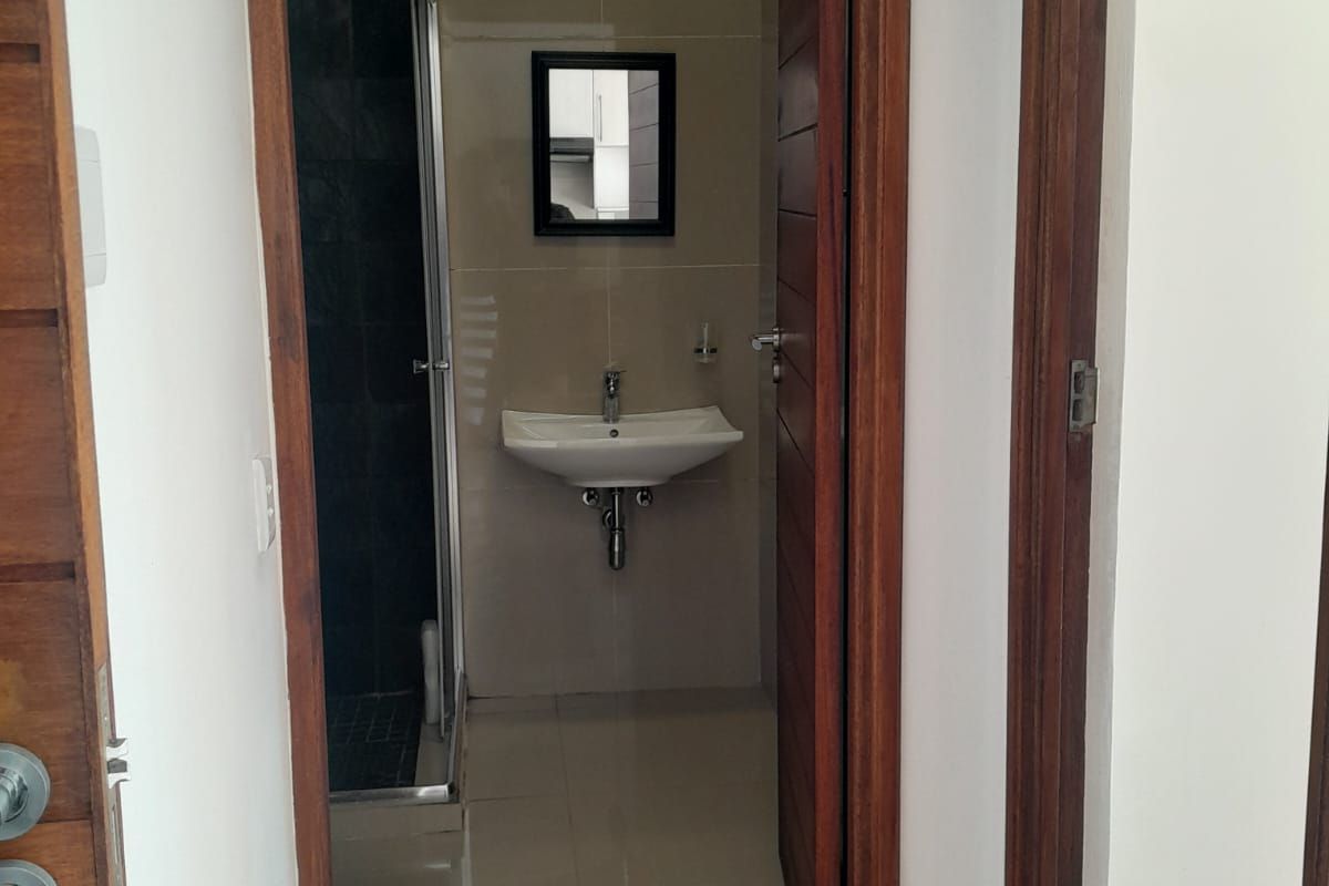 A view through a doorway into a compact bathroom featuring a wall-mounted basin, tiled flooring, and a shower enclosure with dark wall tiles.