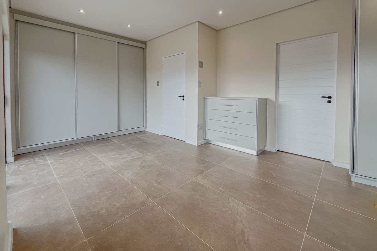 A bright, neutral bedroom featuring large format floor tiles, built-in sliding door wardrobes, and recessed ceiling downlights.