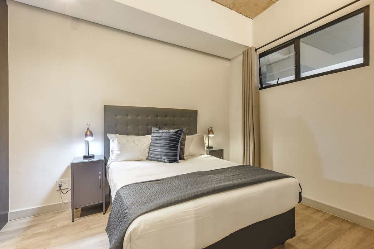 A modern bedroom featuring light wood-look flooring, neutral walls, and a high-level window with black framing.
