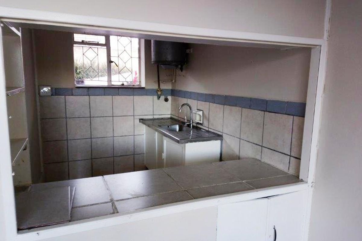 A compact kitchen area featuring a tiled countertop, stainless steel sink, and built-in cabinetry, viewed through a service hatch from the adjacent living space.