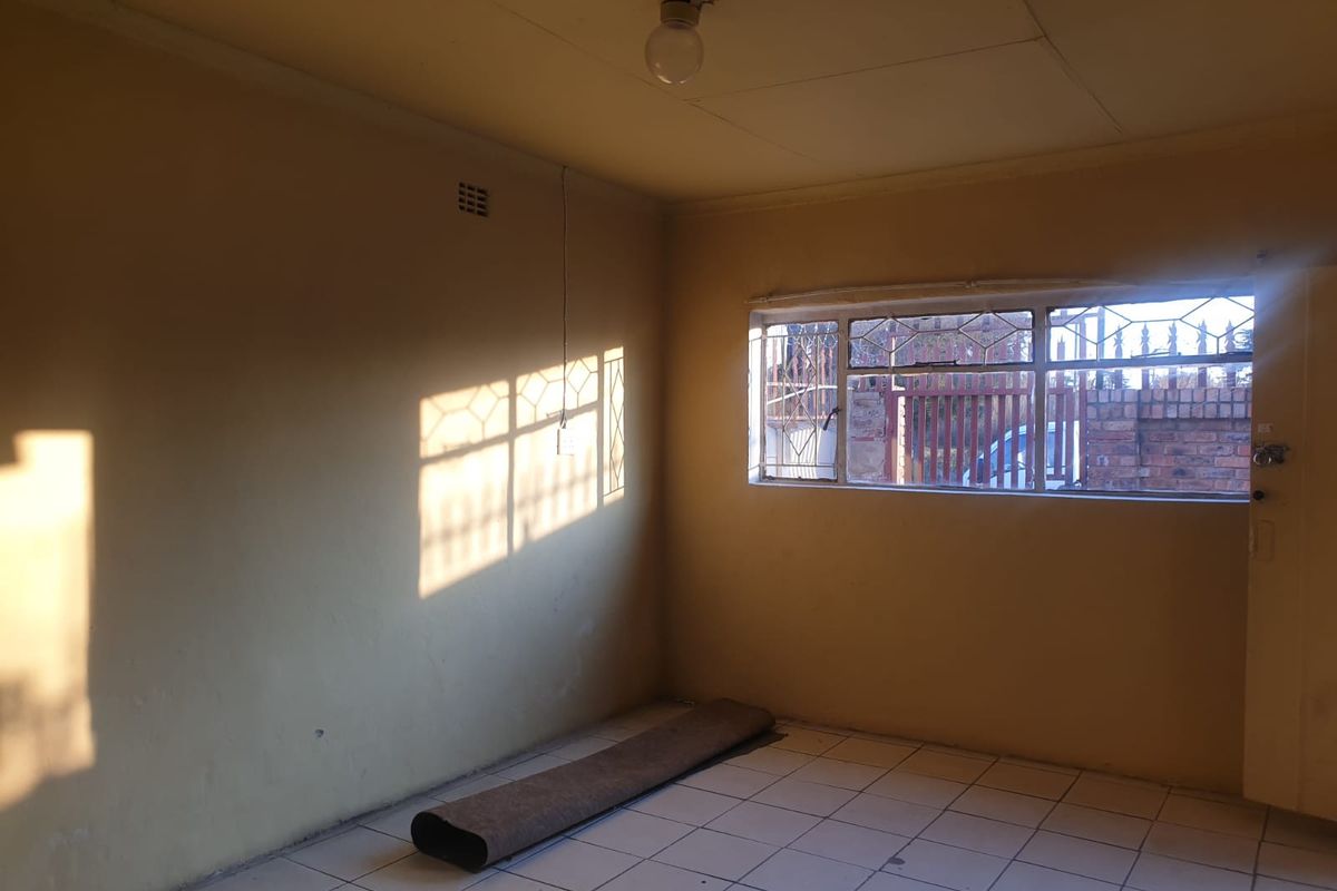 An empty living room featuring tiled flooring, large windows with security bars, and basic lighting fixtures, offering a blank canvas for renovation.