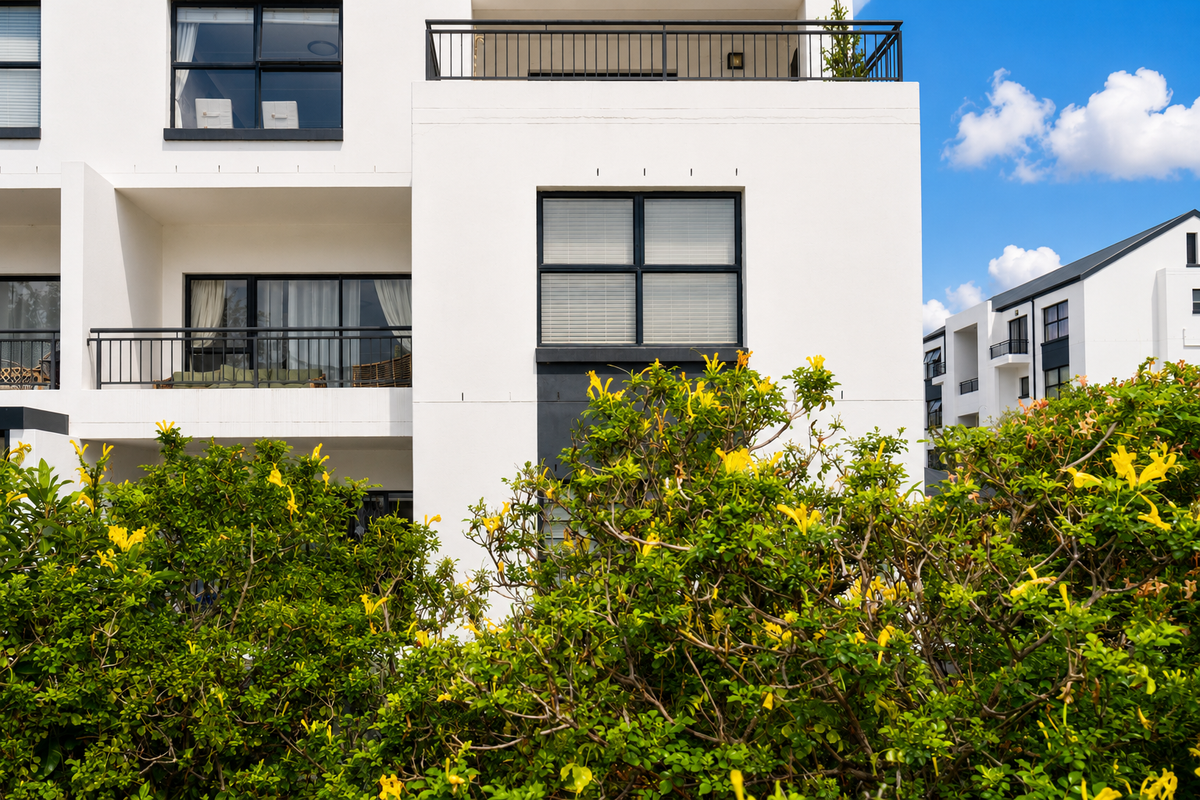 A modern apartment building exterior featuring white facade, black window frames, and private balconies with metal railings, surrounded by lush landscaping.