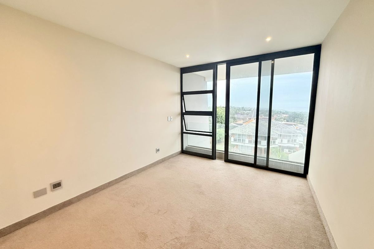 A bright, empty bedroom featuring neutral carpeting, recessed ceiling lighting, and large floor-to-ceiling sliding glass doors providing access to a balcony with neighborhood views.