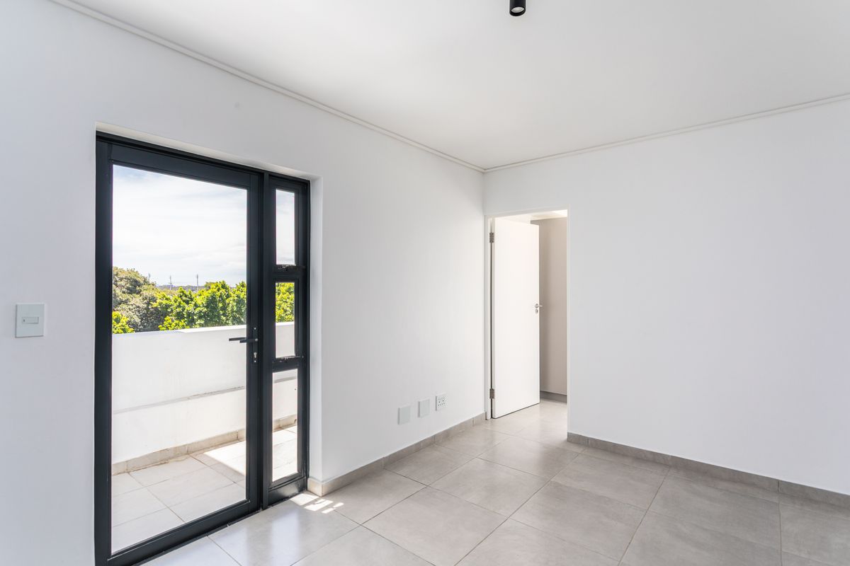 A bright, empty bedroom featuring neutral tiled flooring, white walls, and a black-framed glass door providing access to a private balcony.