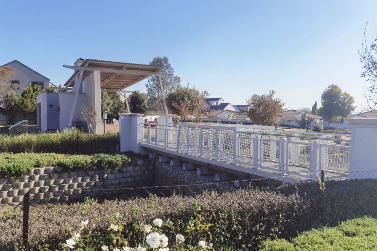 A well-maintained pedestrian bridge with white metal railings leading to a covered outdoor entertainment area within a landscaped residential estate.