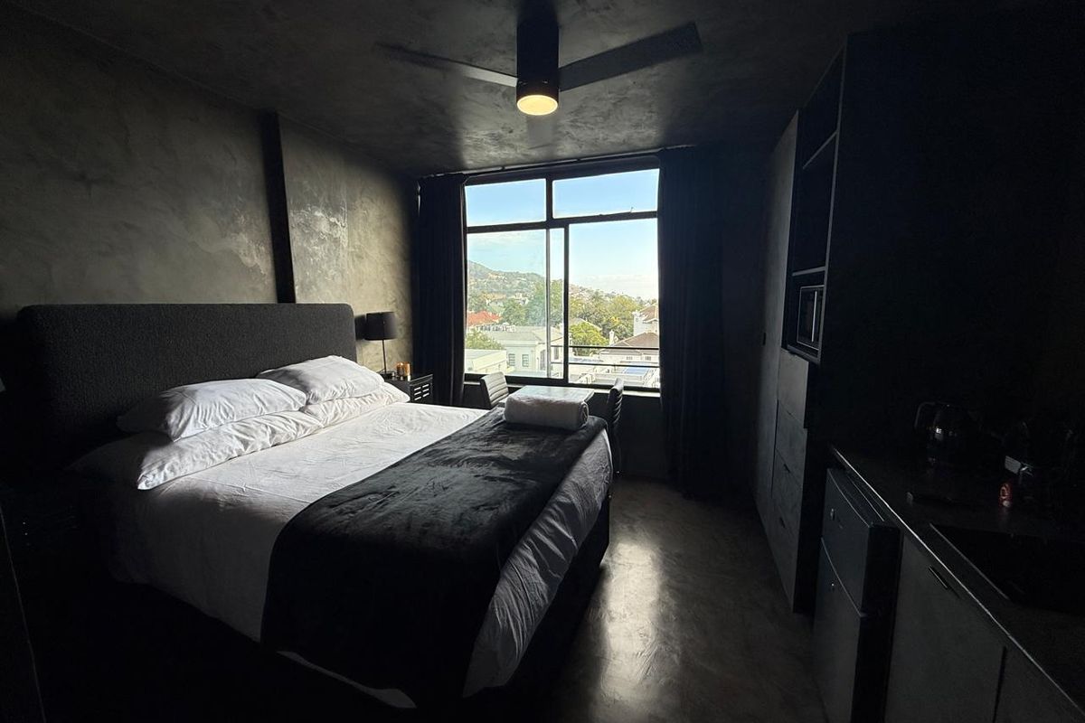 Modern bedroom featuring dark concrete-style walls, polished concrete flooring, a ceiling fan, and large windows offering an elevated urban view.