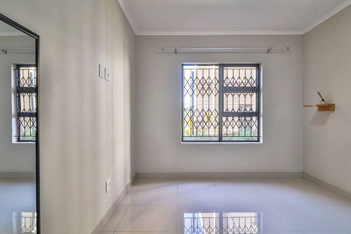 A bright, empty bedroom featuring neutral tiled flooring, clean walls, and a window fitted with security bars and a curtain rod.