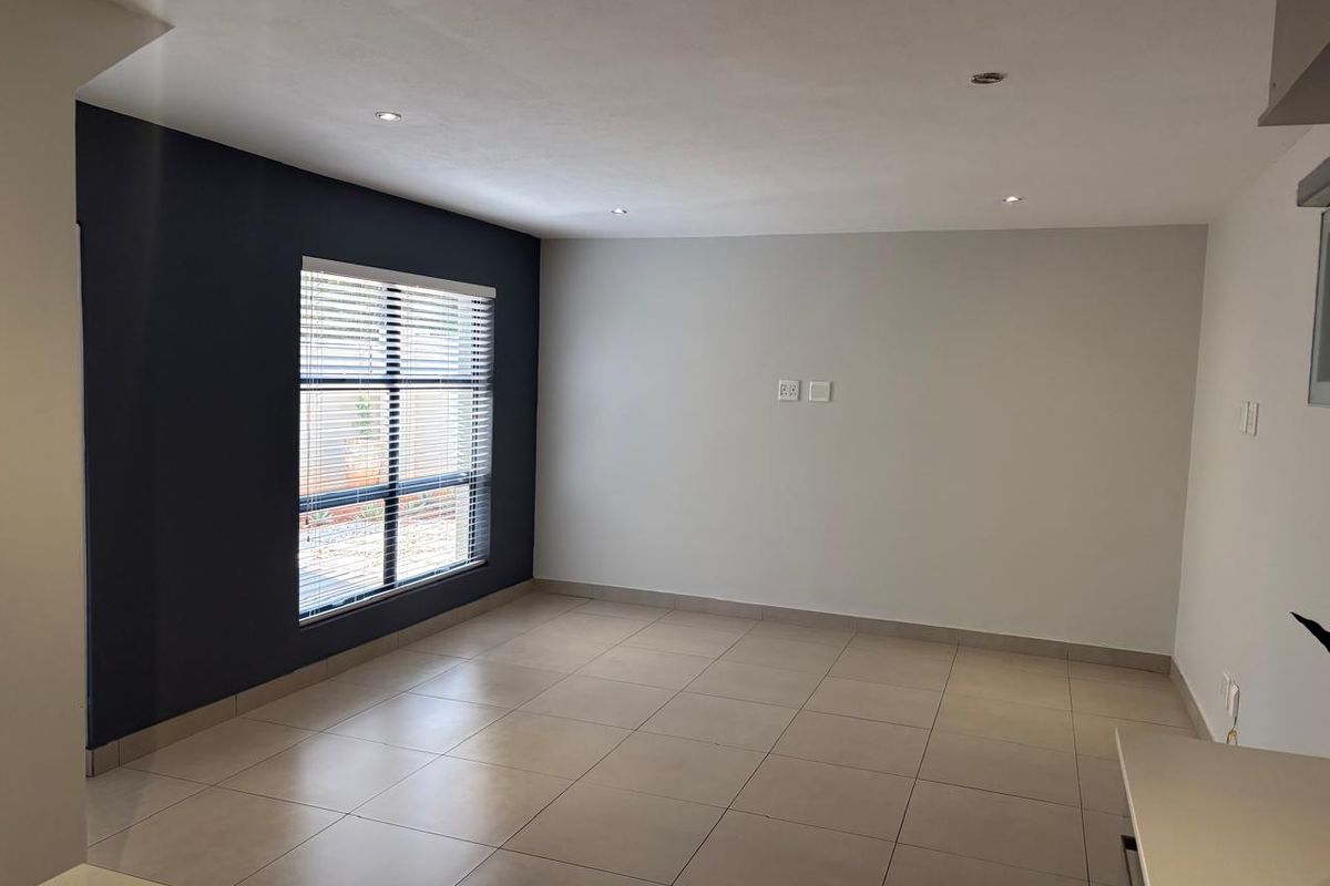 A bright, empty living area featuring neutral floor tiles, recessed ceiling lighting, and a striking dark accent wall with a fitted window blind.