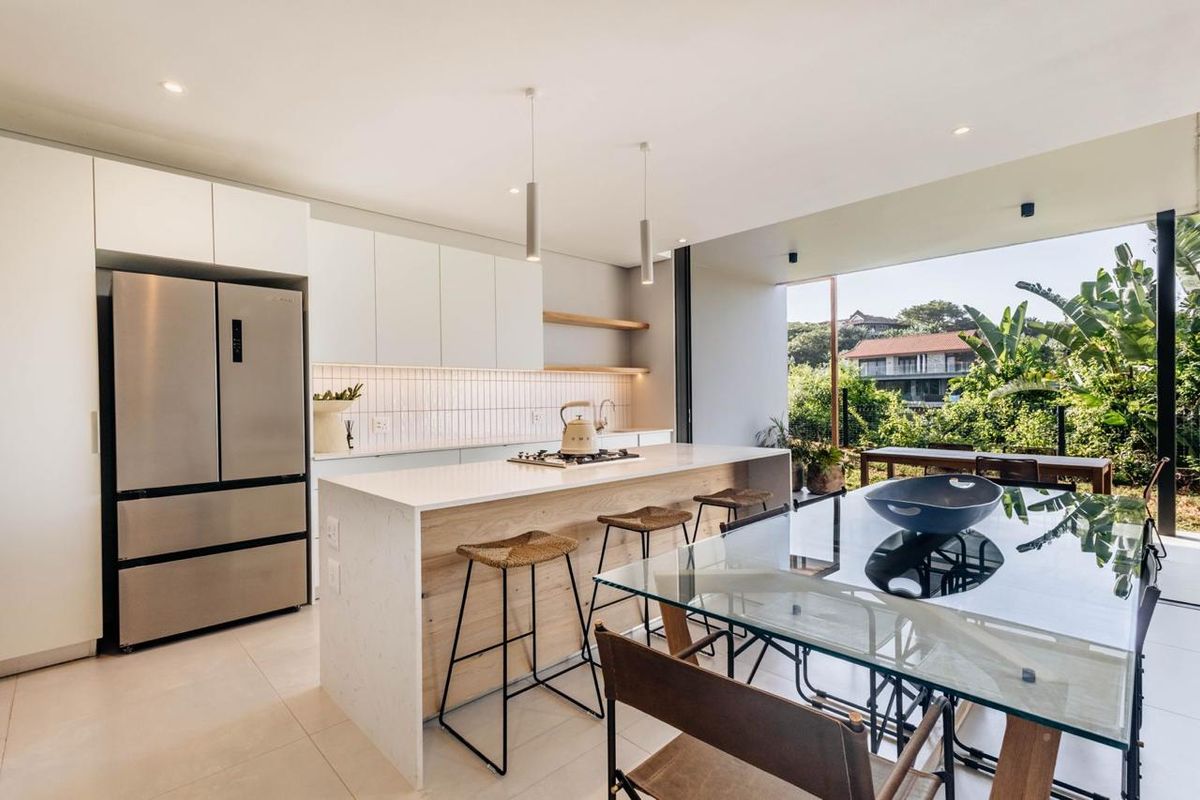 Modern open-plan kitchen and dining area featuring a stone-topped island, sleek cabinetry, and large sliding doors opening to a lush outdoor view.