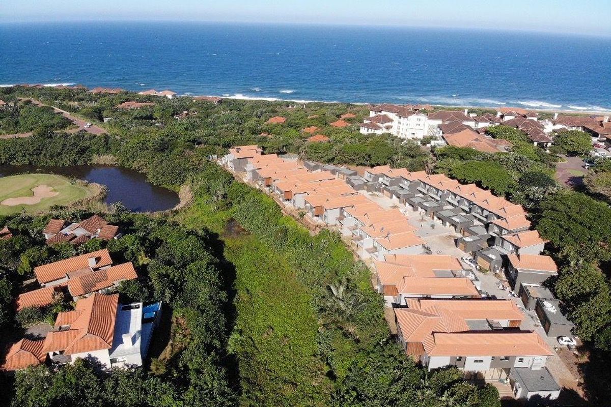 An aerial view of a coastal townhouse estate featuring terracotta-tiled roofs, surrounded by lush greenery and proximity to the ocean.