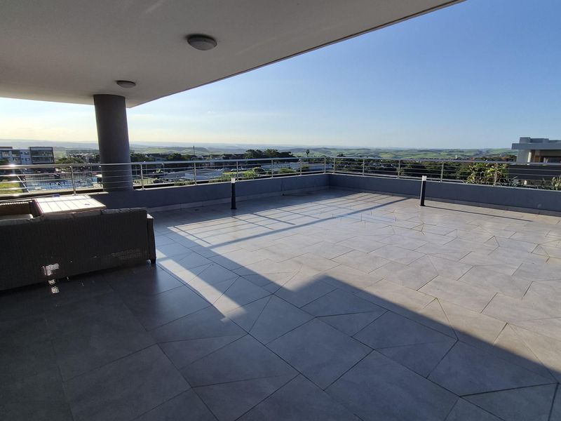 A spacious covered balcony featuring modern tiled flooring, stainless steel railings, and expansive panoramic views of the surrounding landscape.