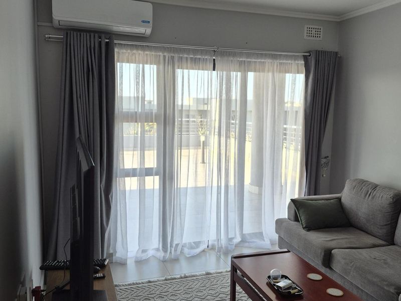 A bright living room featuring large sliding glass doors leading to a balcony, equipped with a wall-mounted air conditioning unit and recessed ceiling lighting.