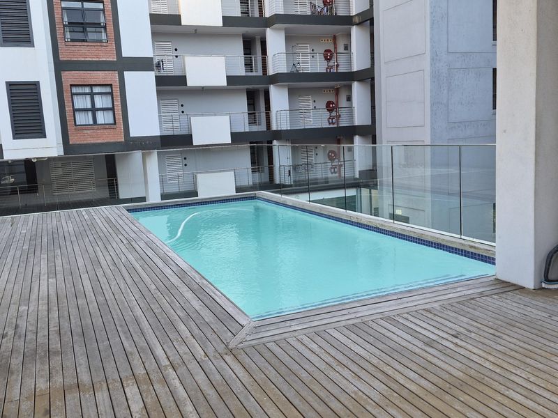 A communal swimming pool area featuring a wooden deck surround and glass balustrades, set within a modern apartment complex.
