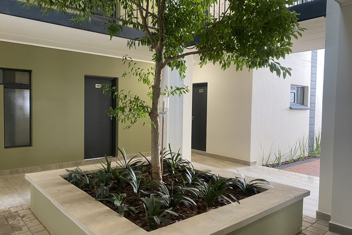 A well-maintained apartment complex courtyard featuring a central raised garden bed with a tree, paved walkways, and access to individual apartment units.