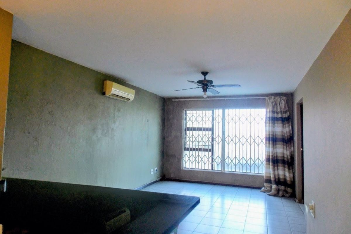 A spacious, empty living area featuring tiled flooring, a ceiling fan, and a wall-mounted air conditioning unit with security bars on the window.