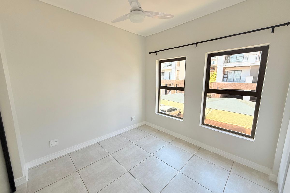 A bright, empty bedroom featuring neutral tiled flooring, a ceiling fan, and large windows with curtain rods, offering a clean and versatile living space.