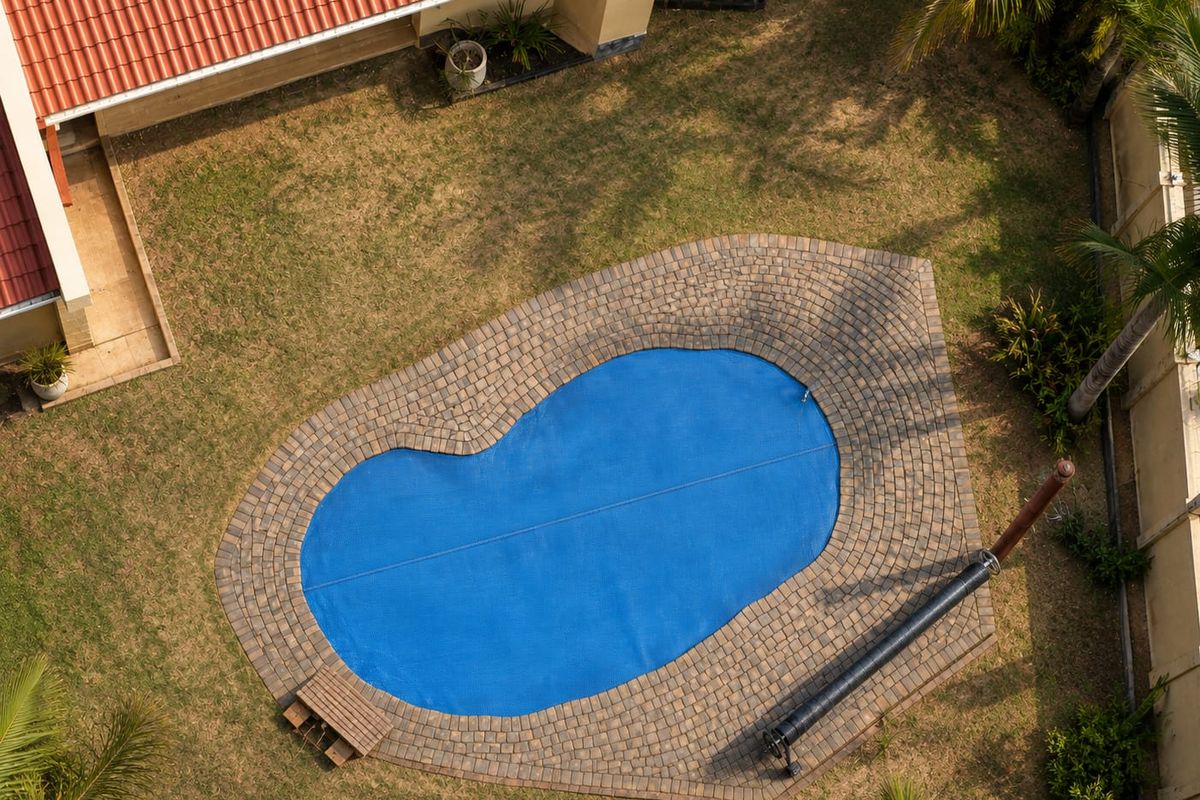 Aerial view of a backyard featuring a kidney-shaped swimming pool with a blue cover, paved surround, and surrounding lawn area.
