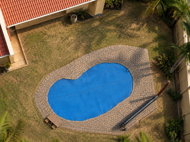 Aerial view of a backyard featuring a kidney-shaped swimming pool with a blue cover, paved surround, and surrounding lawn area.