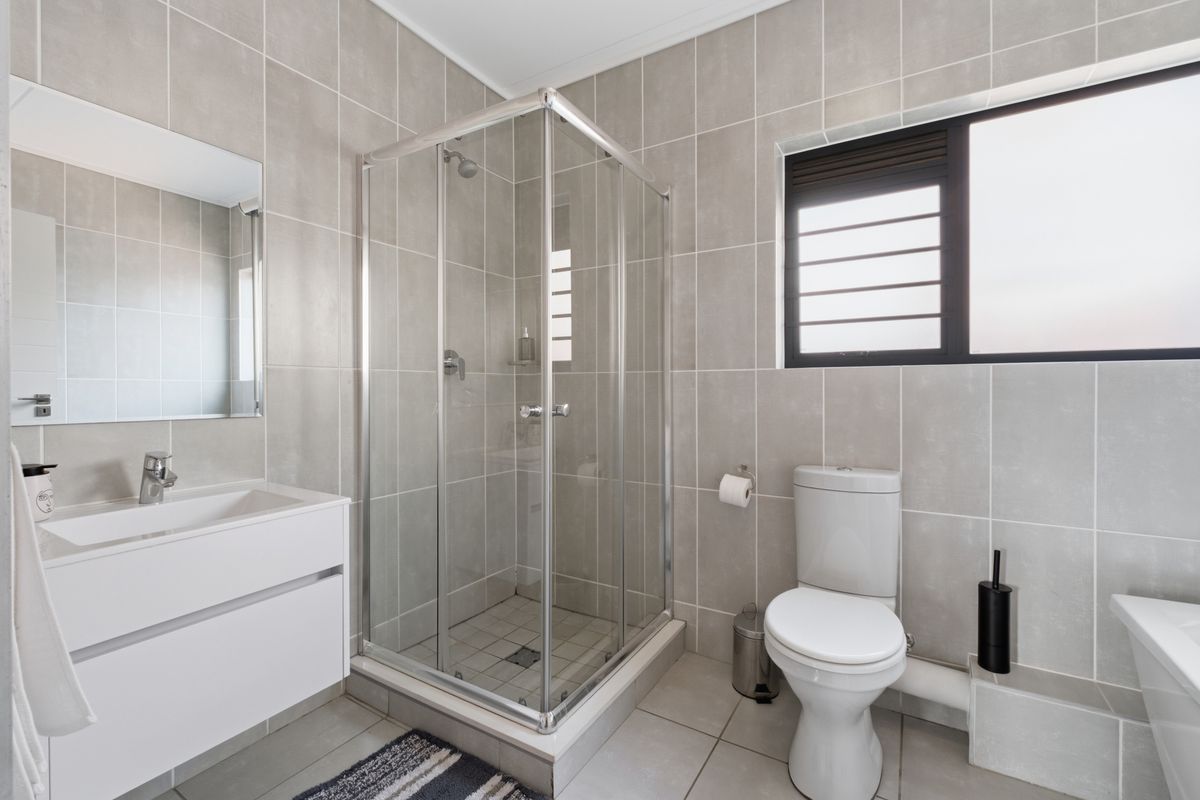 Modern bathroom featuring a glass-enclosed corner shower, wall-mounted vanity with basin, and a white toilet, finished with neutral grey wall and floor tiles.