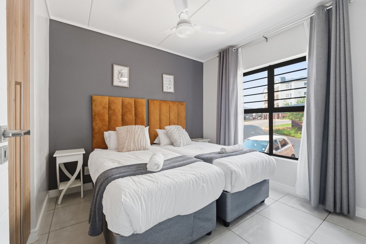 A well-lit bedroom featuring tiled flooring, a ceiling fan, and a large window with security bars, finished with a modern grey accent wall.